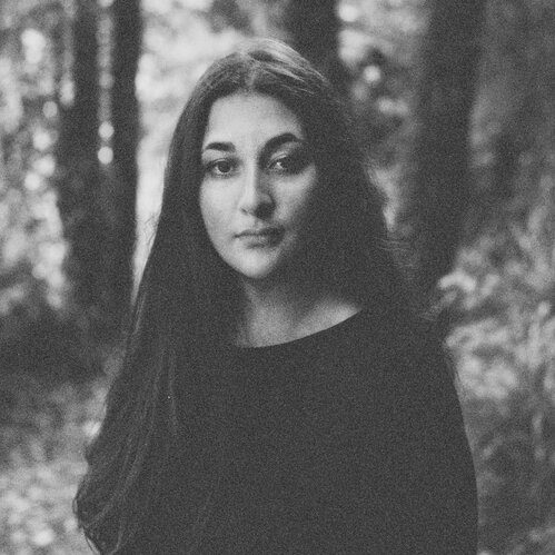 A black and white photo of Lydia Hounat, a British-Algerian woman with long dark hair, wearing a black top. She is standing in some woodland, and looking straight at the camera.